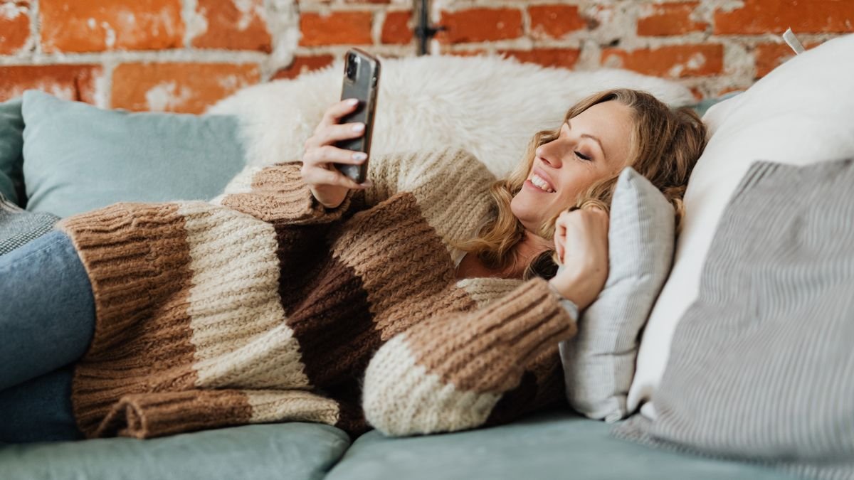Woman in a knitted sweater smiling at her phone on the sofa